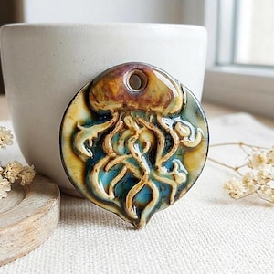 May include: A circular ceramic pendant featuring a raised, embossed jellyfish design. The piece displays a glossy glaze with blended shades of deep blue, teal, and warm brown. A single hole is positioned at the top for hanging or stringing.