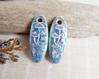 May include: Two oval ceramic pendants featuring a mushroom design. The pendants are light blue with a glossy finish. The mushroom design is in shades of purple and white, with blue accents. Each pendant has a hole at the top for stringing.