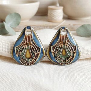 May include: Two triangular ceramic pendants featuring a glossy finish with a blue, green, and brown color palette. Each piece displays a central teardrop shape surrounded by intricate, raised line patterns and includes a circular hole at the top.