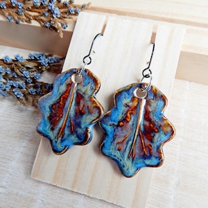 May include: A pair of ceramic leaf-shaped earrings. The earrings feature a blue, brown, and cream color palette with a glossy finish. The earrings are suspended from black metal ear wires. The earrings are displayed on a wooden stand.