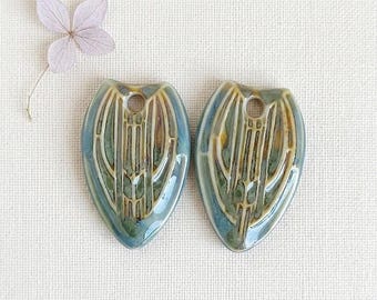 May include: Two ceramic pendant beads, teardrop-shaped with a hole at the top. Glazed in blue, green, and brown, they feature a vertical linear design. A dried lavender flower is in the upper left corner. Measurements are in centimetres.