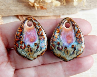 May include: Two ceramic teardrop pendants with a hole at the top. The pendants display a mix of pink, orange, blue, and brown hues. The surface has a glossy finish, and the design appears abstract and organic. Each pendant is approximately 3 cm in length.