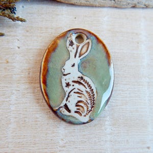 May include: Oval ceramic pendant with a white rabbit design. The pendant has a hole at the top for hanging. The background is a light green with brown and gold edges. The rabbit is detailed with etched lines.