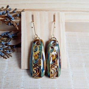 Könnte beinhalten: Ein Paar Keramikohrringe mit einem blauen, braunen und goldenen Design. Die Ohrringe sind rechteckig mit abgerundeten Ecken und haben einen goldenen Haken.