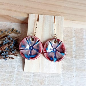 May include: A pair of ceramic earrings with a circular design. The earrings feature a red, white, and blue color scheme with a floral pattern. The earrings are suspended from gold-colored hooks and are displayed on a wooden stand.