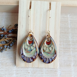 Puede incluir: Un par de pendientes de cerámica en forma de lágrima con un diseño en capas. Los pendientes son una mezcla de marrón, verde y morado con detalles dorados. Los pendientes cuelgan de ganchos dorados.