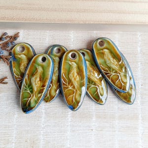 May include: A collection of seven ceramic pendants. Each pendant is oval-shaped with a bird design in shades of green, brown, and white. The edges are outlined in blue. Each pendant has a hole at the top for stringing.