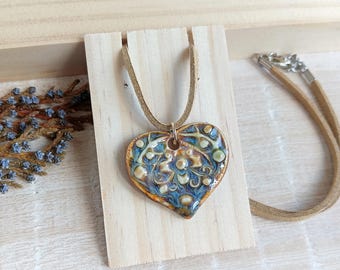 May include: A heart-shaped ceramic pendant with a brown leather cord. The pendant features a blue, brown, and white glazed design with raised details. The necklace is displayed on a wooden stand.