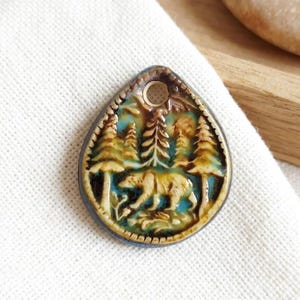 May include: A teardrop-shaped ceramic pendant featuring a relief design of a bear walking through a forest of pine trees. The piece is glazed in shades of blue, cream, and brown, with a circular hole at the top for suspension on a cord or chain.