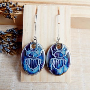 Può includere: Un paio di orecchini ovali con un design a scarabeo. Gli orecchini presentano una smaltatura blu e viola con un contorno di scarabeo bianco. Sono appesi a ganci color argento e sono esposti su uno sfondo di legno.