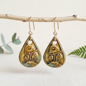 Puede incluir: Un par de pendientes de cerámica en forma de lágrima con un diseño en relieve de setas con un motivo de ojo. Los pendientes tienen un acabado esmaltado brillante en tonos verde oliva y marrón, suspendidos de ganchos de metal dorado.