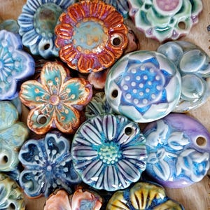 Könnte beinhalten: Eine Sammlung farbenfroher Keramikblumenperlen in verschiedenen Formen und Größen. Die Perlen haben eine glänzende Oberfläche und eine Reihe von Farben, darunter Blau, Türkis, Orange und Lila. Jede Perle hat ein Loch zum Auffädeln, ideal für Schmuck oder andere Bastelarbeiten.