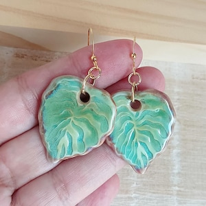 Puede incluir: Un par de pendientes con forma de hoja. Cada pendiente presenta un diseño detallado pintado a mano con tonos verdes y azules, y un gancho dorado. Los pendientes son de cerámica.