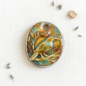 May include: An oval ceramic pendant featuring a relief carving of a bird perched on a branch. The surface has a glossy glaze with blended tones of turquoise, teal, and warm brown. A circular hole is positioned at the top for hanging purposes.