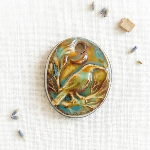 May include: An oval ceramic pendant featuring a relief carving of a bird perched on a branch. The surface has a glossy glaze with blended tones of turquoise, teal, and warm brown. A circular hole is positioned at the top for hanging purposes.