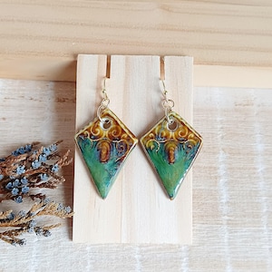 Könnte beinhalten: Ein Paar rautenförmige Keramikohrringe mit goldfarbenen Haken. Die Ohrringe haben eine grüne und braune Glasur mit einem dekorativen Muster. Die Ohrringe werden auf einem Holzständer präsentiert.