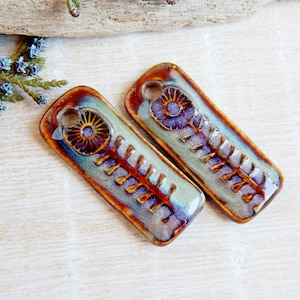 May include: Two ceramic rectangular pendants with a floral design. The pendants are glazed in shades of blue, green, and brown with a purple flower design.