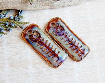 May include: Two ceramic rectangular pendants with a floral design. The pendants are glazed in shades of blue, green, and brown with a purple flower design.
