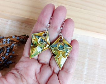 May include: A pair of handmade ceramic earrings with a geometric design. The earrings feature a diamond shape with a central blue accent, surrounded by green, yellow, and white glaze. The earrings are suspended from silver-colored hooks.