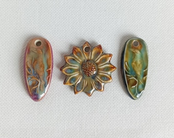 May include: Three ceramic pendants with glossy glazes. The left and right pendants are oval-shaped with embossed bird designs in blue, brown, and green. The center pendant is a flower shape with a textured brown center and brown petals.