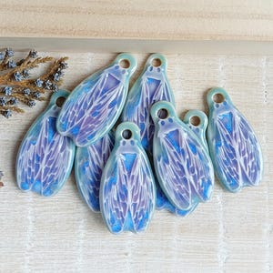 Op de afbeelding: Een set van zes keramische cicada-charms met iriserende blauwe en paarse vleugels. De charms zijn geglazuurd met een lichtblauwe en groene glazuur.