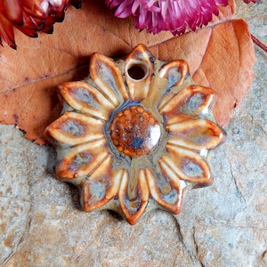 Puede incluir: Un adorno de cerámica en forma de girasol con un esmalte marrón y blanco. El girasol tiene un centro elevado y pétalos. El adorno tiene un agujero en la parte superior para colgarlo.
