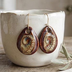 Può includere: Un paio di orecchini in ceramica a forma di goccia con una smaltatura lucida marrone scuro e un motivo centrale in rilievo a forma di foglia di felce color crema. Gli orecchini pendono da sottili ganci in metallo color oro.
