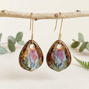 Puede incluir: Un par de pendientes de cerámica en forma de lágrima con un esmalte reactivo que presenta patrones de remolinos en marrón oscuro, magenta vibrante y azul suave. Los pendientes cuelgan de finos ganchos de metal dorado curvados.
