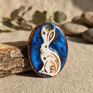 Op de afbeelding: Ovale keramische hanger met een wit konijnontwerp op een blauwe achtergrond. De hanger heeft een bruine rand en een gat om op te hangen. Het konijn is gedetailleerd met steraccenten.