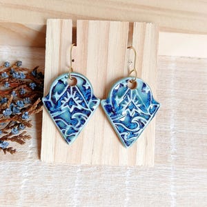 Puede incluir: Un par de pendientes de cerámica con un diseño azul y blanco. Los pendientes tienen forma de diamante con un patrón decorativo y ganchos dorados. Los pendientes se exhiben en un soporte de madera.