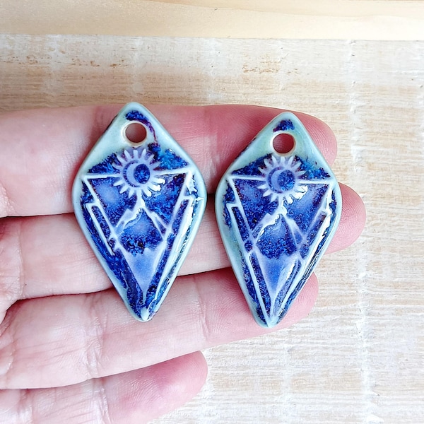 May include: Two blue and white ceramic pendants with a diamond shape. Each pendant features a mountain and sun design. The pendants are approximately 1.5 inches long and have a hole at the top for hanging.