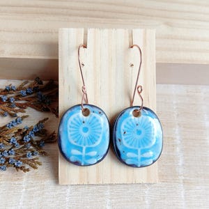 May include: A pair of blue and white ceramic earrings with a floral design. The earrings are hanging from a wooden display stand.