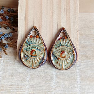 Könnte beinhalten: Ein Paar Keramikohrringe in Tropfenform mit einem Augenmotiv. Die Ohrringe sind braun, grün und weiß mit einer goldenen Glasur.