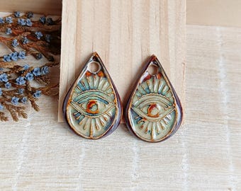 May include: A pair of ceramic earrings in the shape of teardrops with an eye design. The earrings are brown, green, and white with a gold glaze.