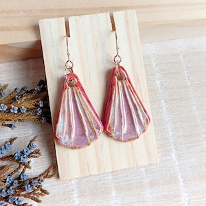 Könnte beinhalten: Ein Paar handgefertigte Ohrringe mit fächerförmigem Design. Die Ohrringe haben ein rosa-weißes Farbschema mit orangefarbenen Akzenten und kupferfarbenen Ohrhaken. Die Ohrringe werden auf einem Holzständer präsentiert.