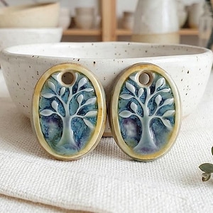 May include: Two oval ceramic pendants featuring a raised white tree design against a blue and purple glazed background. Each pendant has a smooth yellow border and a circular hole at the top for hanging, showcasing a glossy, multi-colored finish.