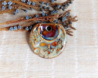 May include: A ceramic bud vase with a round shape and a central hole. The vase features a floral design with blue, orange, and brown hues. The background includes dried branches with small blue flowers.