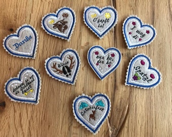 Puede incluir: Nueve adornos de fieltro en forma de corazón con costuras azules y varias frases alemanas bordadas en ellos, incluyendo "O'zapft is", "Wer ko, der ko", "I mog di", "Spatzl", "Wiesn", "Dohoam is wo de Schlossl basst", "Danke" y "Oktoberfest".