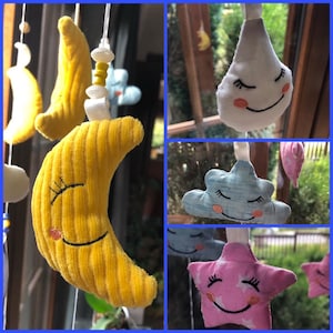 Könnte beinhalten: Ein Baby-Mobile mit Plüsch-Himmelskörpern. Das Mobile enthält einen gelben Halbmond, eine weiße Wolke, eine blaue Wolke und einen rosa Stern, jeweils mit gestickten Gesichtern. Die Formen sind an weißen Schnüren aufgehängt.