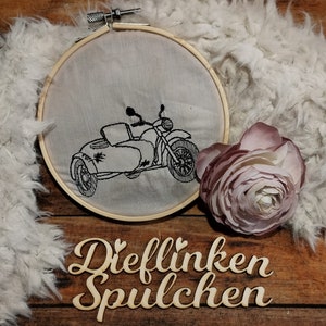 Könnte beinhalten: Ein gesticktes Bild in Schwarzweiß von einem Motorrad mit Beiwagen auf einem weißen Stoff, der über einen hölzernen Stickrahmen gespannt ist. Der Rahmen liegt auf einer Holzoberfläche mit einer rosa Blume und einem Holzschild mit der Aufschrift "Dieflinken Spulchen".