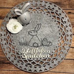 Könnte beinhalten: Ein grauer Filz-Tischset mit einem ausgeschnittenen Design zeigt ein schwarzes gesticktes Häschen und die Worte "Dief linken Spulchen".