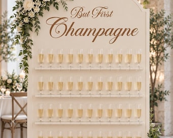 May include: A tall, arched, cream-coloured wooden display wall featuring four horizontal shelves holding rows of clear champagne flutes filled with a golden liquid. The text "But First Champagne" is printed in brown script at the top centre.