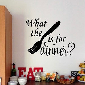 May include: A kitchen with a white wall and a black vinyl decal that says "What the fork is for dinner?" with a fork silhouette. The kitchen has wooden cabinets, a microwave, and a counter with fruit and other food items.