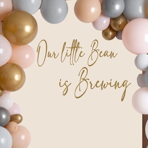 May include: A rectangular backdrop featuring the text Our little Bean is Brewing in gold script, framed by a balloon arch in shades of metallic gold, matte grey, soft blush pink, and white, creating a decorative display for an event.