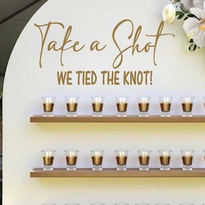 May include: A white wedding display with three wooden shelves holding shot glasses filled with a dark liquid. The text "Take a Shot WE TIED THE KNOT!" is written in gold script. Pink and white flowers decorate the display.