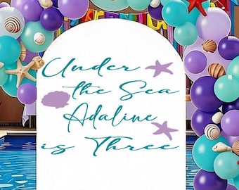 May include: An arched backdrop featuring the text Under the Sea Adaline is Three in teal script, surrounded by a balloon garland in shades of purple, teal, and blue, accented with starfish and seashells, positioned by a swimming pool.