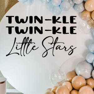 May include: White round sign with black text that reads "Twin-kle Twin-kle Little Stars". The sign is surrounded by blue and beige balloons.