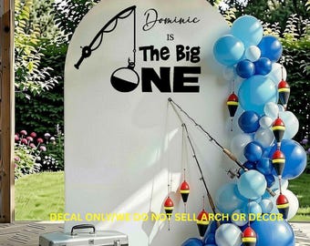 Puede incluir: Un fondo arqueado blanco con letras negras que dicen Dominic IS The Big ONE y un gráfico de caña de pescar. Un grupo de globos azules y adornos de boyas de pesca están sujetos al lado derecho del arco.