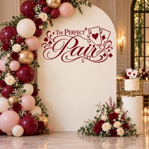 May include: A large arched backdrop featuring a balloon garland in burgundy, blush pink, cream, and gold. The center displays the maroon text The Perfect Pair with playing card graphics. A small cake sits on a fluted pedestal with floral accents.