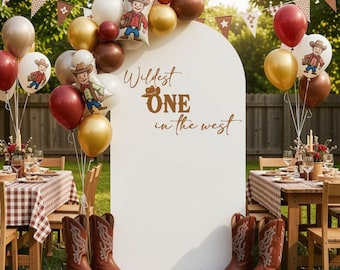 May include: A white arched backdrop featuring the brown text Wildest ONE in the west, surrounded by a balloon garland in red, gold, white, and brown with cowboy illustrations. Two pairs of brown leather cowboy boots sit on the grass below.